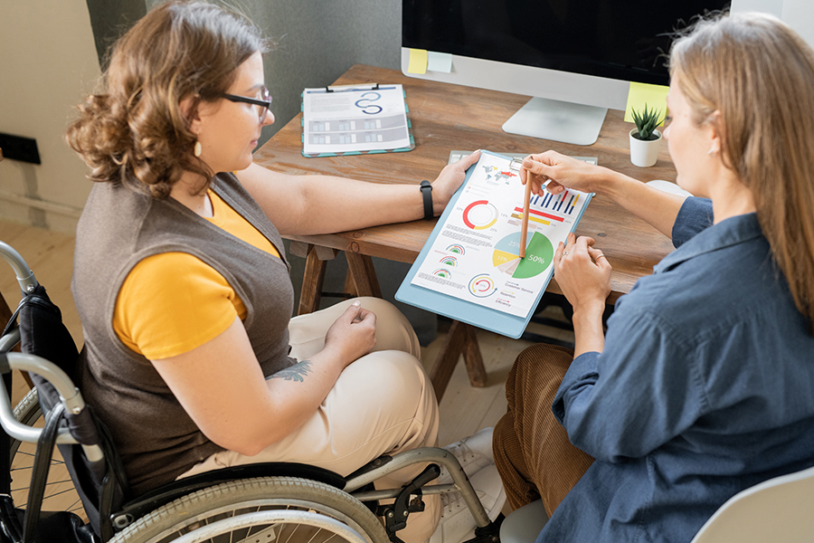 Dos mujeres revisan juntas un informe con gráficos de colores sobre grados de discapacidad, una de ellas en silla de ruedas, en un entorno de oficina.